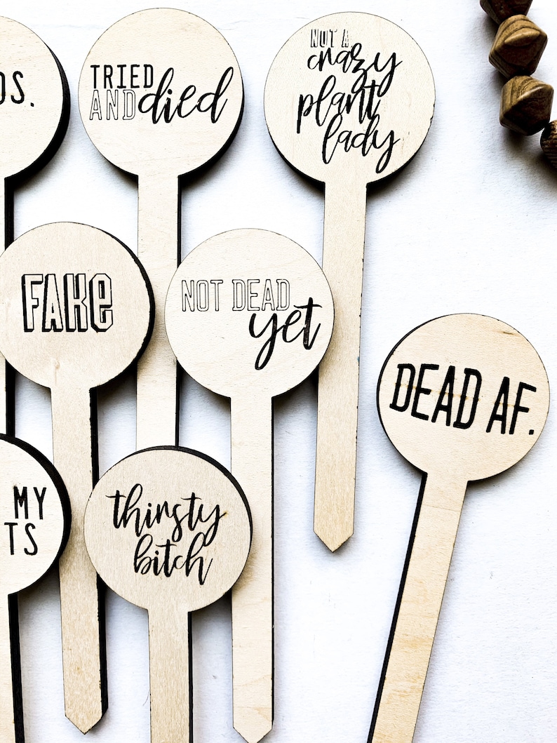 May include: Set of 8 wooden plant stakes with funny sayings. The stakes are round and have black text on a natural wood background. The sayings include "Tried and Died", "Not a Crazy Plant Lady", "Fake", "Not Dead Yet", "Dead AF", "Thirsty Bitch", "My T's" and "DS."