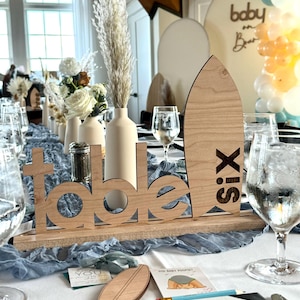 May include: A wooden table number sign with the word "table" and the number "six" on a surfboard shape. The sign is on a table with a blue tablecloth and white flowers in vases.