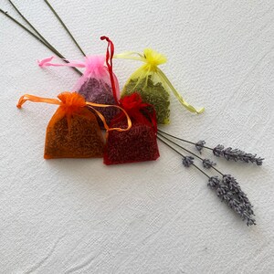 K&ouml;nnte beinhalten: Vier kleine Stoffbeutel gef&uuml;llt mit getrockneten Lavendelbl&uuml;ten. Die Beutel sind in den Farben Rosa, Orange, Rot und Gelb. Die Beutel liegen auf einer wei&szlig;en Oberfl&auml;che mit einem Lavendelzweig.