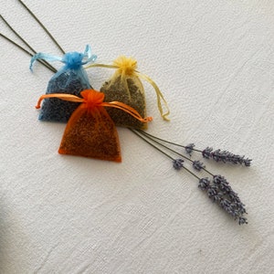 K&ouml;nnte beinhalten: Drei kleine Stoffbeutel gef&uuml;llt mit getrockneten Lavendelbl&uuml;ten. Die Beutel sind blau, gelb und orange. Ein Lavendelzweig befindet sich im Hintergrund.