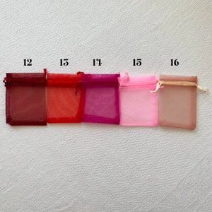 K&ouml;nnte beinhalten: F&uuml;nf kleine, quadratische, durchsichtige Stoffbeutel in verschiedenen Farben. Die Beutel sind von links nach rechts angeordnet: Burgunderrot, Rot, Rosa, Hellrosa und Beige. Jeder Beutel hat einen Kordelverschluss und ein kleines Etikett mit einer Nummer darauf. Die Nummern sind 12, 15, 17, 15 und 16.
