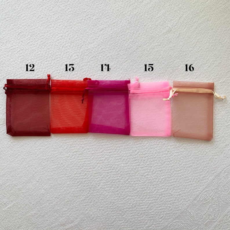 K&ouml;nnte beinhalten: F&uuml;nf kleine, quadratische, durchsichtige Stoffbeutel in verschiedenen Farben. Die Beutel sind von links nach rechts angeordnet: Burgunderrot, Rot, Rosa, Hellrosa und Beige. Jeder Beutel hat einen Kordelverschluss und ein kleines Etikett mit einer Nummer darauf. Die Nummern sind 12, 15, 17, 15 und 16.