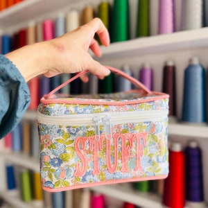 May include: A floral print cosmetic bag with the word "STUFF" embroidered in pink. The bag has a pink handle and a white zipper. The background features shelves filled with colorful spools of thread.