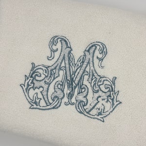 May include: A cream-colored terry cloth towel featuring an embroidered letter M in a dark blue, ornate, swirling script font. The embroidery is centered on the fabric, showcasing detailed line work and decorative flourishes within the letter.