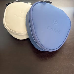 May include: Two Therabody-branded, zippered cases. One is a light cream colour, the other is a muted blue. Both have a rounded, square shape and a zip closure. The Therabody logo and brand name are printed on the front of each case.