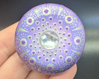 Pode incluir: Uma pedra pintada de roxo e prata com um desenho de mandala e um strass transparente no centro.