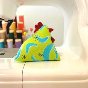 May include: A handmade chicken-shaped pin cushion with a lime green background and blue boomerang shapes. The chicken has a red comb and two pins are inserted. The pin cushion is on a white sewing machine.