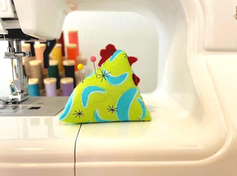 May include: A handmade chicken-shaped pin cushion with a lime green background and blue boomerang shapes. The chicken has a red comb and two pins are inserted. The pin cushion is on a white sewing machine.
