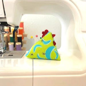 May include: A lime green chicken-shaped pin cushion with blue crescent and star patterns, and a red comb. Several sewing pins are inserted into the cushion. The pin cushion is on a white sewing machine.
