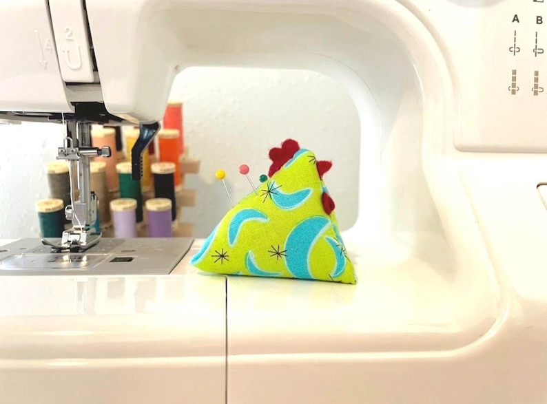 May include: A lime green chicken-shaped pin cushion with blue crescent and star patterns, and a red comb. Several sewing pins are inserted into the cushion. The pin cushion is on a white sewing machine.