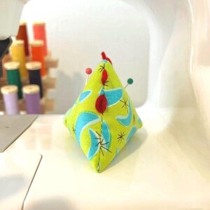 May include: A handmade, diamond-shaped pin cushion with a lime green background and a retro blue and white pattern. Several pins are inserted into the cushion, and a small red felt accent is on top. Sewing notions are in the background.