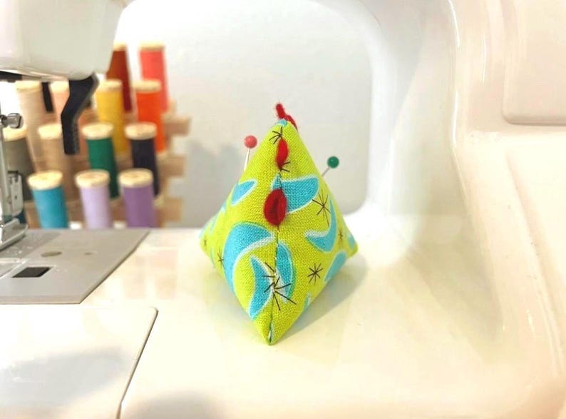 May include: A handmade, diamond-shaped pin cushion with a lime green background and a retro blue and white pattern. Several pins are inserted into the cushion, and a small red felt accent is on top. Sewing notions are in the background.