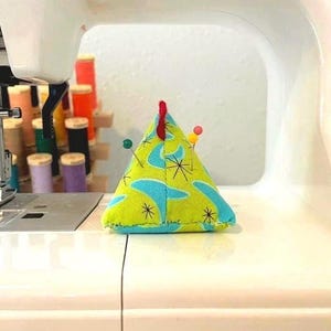 May include: A triangular pin cushion with a retro design in shades of green and blue, adorned with several pins. The cushion sits on a sewing machine, suggesting its use in sewing and crafting projects.