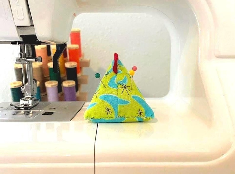 May include: A triangular pin cushion with a retro design in shades of green and blue, adorned with several pins. The cushion sits on a sewing machine, suggesting its use in sewing and crafting projects.