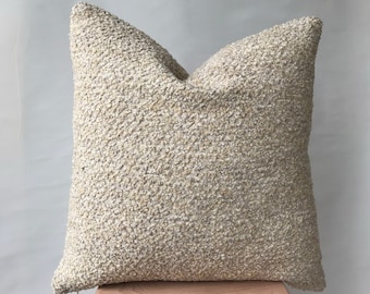 Puede incluir: Una almohada de sofá de color crema con una tela texturizada y esponjosa. La almohada está sentada en un taburete de madera.