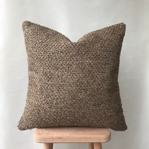 May include: A brown and beige textured throw pillow with a square shape. The pillow is sitting on a wooden stool.