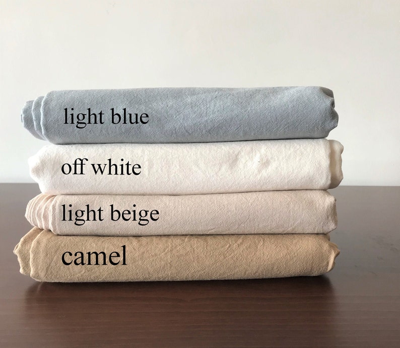 May include: Four folded fabric samples in light blue, off-white, light beige, and camel colors are stacked on a dark brown surface. The colors are labeled with text: "light blue", "off white", "light beige", and "camel".