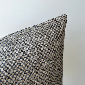 May include: Close-up of a woven pillow corner. The pillow features a textured pattern in shades of beige and navy blue. The fabric appears to be tightly woven, creating a geometric design. The pillow is likely for home decor.