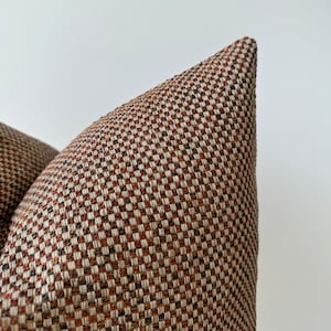 Può includere: Primo piano di un cuscino a quadri marroni e beige con un sottile motivo geometrico. Il cuscino è realizzato in tessuto tessuto con una texture morbida.
