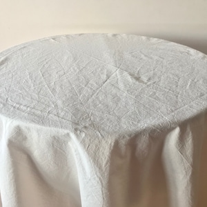 May include: A white, round tablecloth draped over a table. The fabric has a textured appearance with visible creases and folds. The table is partially visible, with a light-colored wall in the background.