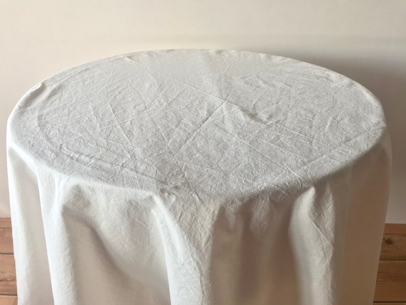 May include: A white, round tablecloth draped over a table. The fabric has a textured appearance with visible creases and folds. The table is partially visible, with a light-colored wall in the background.