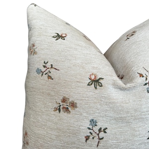 May include: Close-up of a beige pillow with a floral pattern. The pillow has a subtle texture and features small, multi-colored flowers.