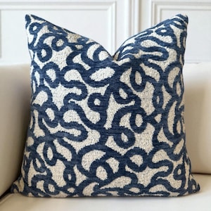 Può includere: Un cuscino decorativo quadrato con un motivo astratto blu navy e bianco sporco. Il cuscino presenta un tessuto strutturato con un design a spirale e ad anello. Il cuscino è posto su un divano di colore chiaro.