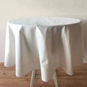 May include: A white, round tablecloth draped over a small table. The fabric has a textured appearance, and the table is supported by a light-colored wooden frame. The tablecloth is made of a soft material.