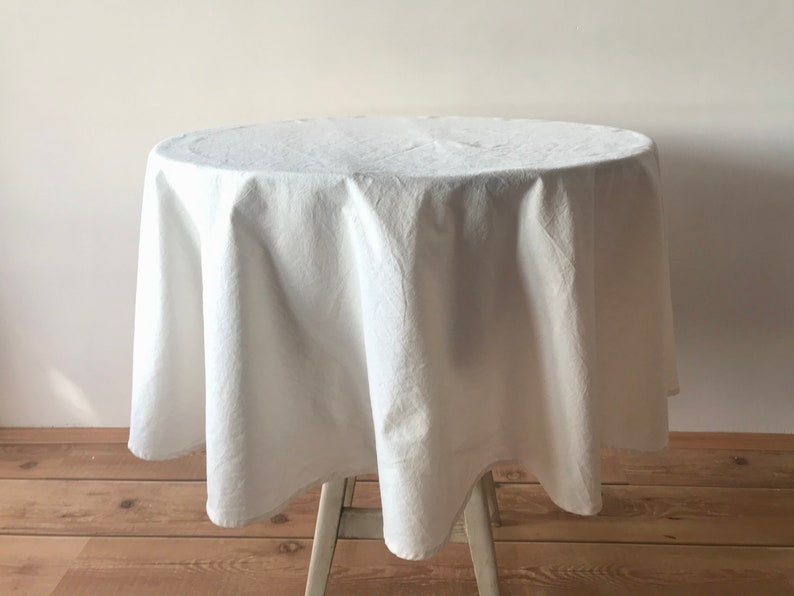May include: A white, round tablecloth draped over a small table. The fabric has a textured appearance, and the table is supported by a light-colored wooden frame. The tablecloth is made of a soft material.