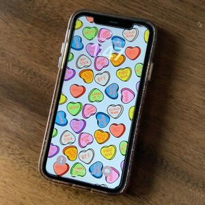 May include: A smartphone with a white background and a pattern of colorful candy hearts with various messages, including "Call Me", "Soul Mate", "BFF", "First Kiss", "Hugs", "Cloud Nine", "Sweet", "Love", "Wow", "38", "Mine", "Hottie", and "You".
