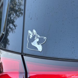May include: A silver and black husky dog decal is affixed to the rear window of a black vehicle. The decal is a side profile of the dog's head, showcasing its distinctive features.