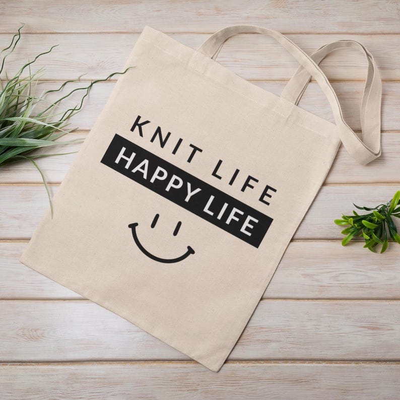 Tote bag with wood plank back drop.  Tote bag says "knit life happy life"