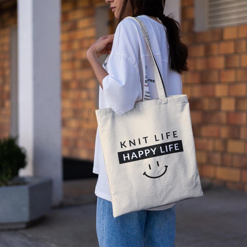 Woman wearing blue jeans with white shirt holding a tote bag that says "Knit Life Happy Life"