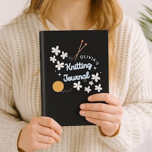 May include: A black journal with the text "Olivia's Knitting Journal" in white script, adorned with white flower and yarn ball illustrations. The journal is held by a person wearing a cream-colored sweater.