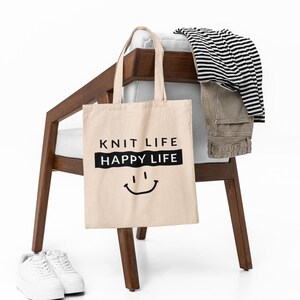 Tote bag hanging on a chair with clothes laid out, bag says "Knit Life Happy Life"