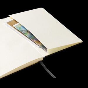 May include: A black hardcover notebook with a cream-colored interior and a black ribbon bookmark. The notebook is open to a blank page with a colorful piece of paper tucked into the pocket.