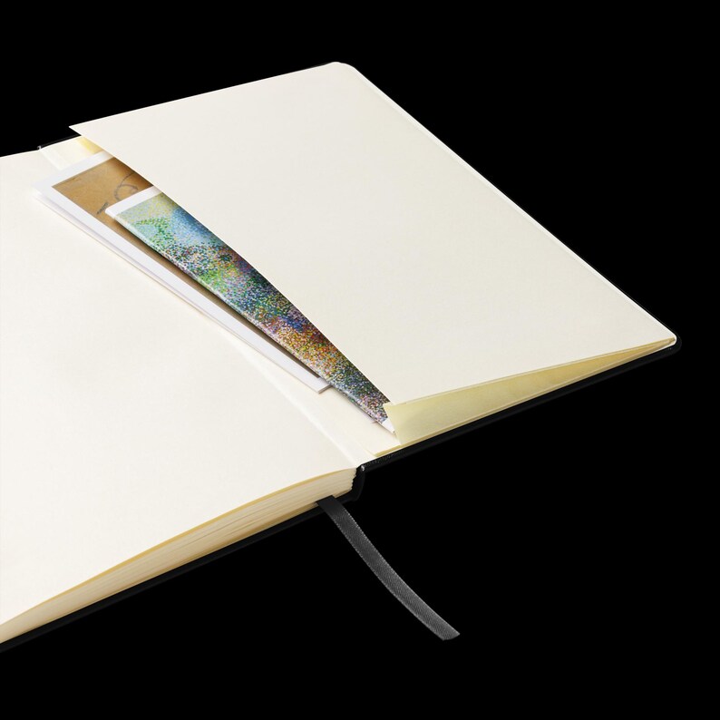 May include: A black hardcover notebook with a cream-colored interior and a black ribbon bookmark. The notebook is open to a blank page with a colorful piece of paper tucked into the pocket.