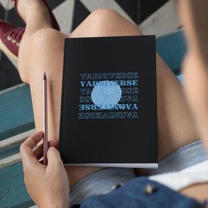 May include: A black notebook with the text "Yarn Universe" repeated in a pattern. A blue yarn ball graphic is centered on the notebook.