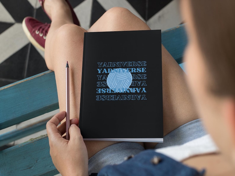 May include: A black notebook with the text "Yarn Universe" repeated in a pattern. A blue yarn ball graphic is centered on the notebook.
