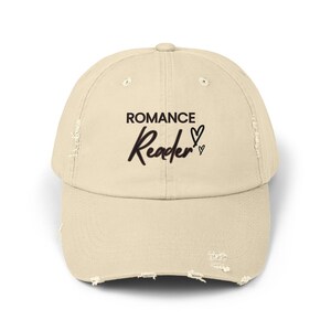 Może przedstawiać: Beżowa, zniszczona czapka z daszkiem z napisem "ROMANCE Reader" w kolorze czarnym z dwoma małymi sercami.