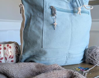 May include: A light blue canvas tote bag with a front zipper pocket and a star-shaped charm. The bag has a drawstring closure and a cream-colored strap. Knitting supplies, including yarn, needles, and stitch markers, are visible.