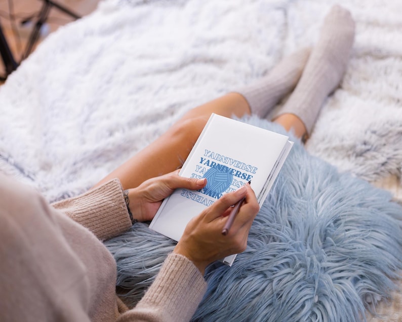 May include: A person sitting on a fluffy blue rug, holding a white notebook with the text "Yarn Universe" written on it. The notebook has a blue yarn ball graphic on the cover.