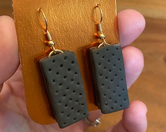 May include: A pair of novelty earrings featuring a dark brown rectangular design with small, evenly spaced holes, resembling a sandwich cookie. The earrings are suspended from gold-colored ear wires and are displayed on a gold-colored card.
