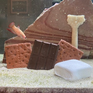 May include: A close-up of a s'mores themed aquarium decoration. The decoration includes a chocolate bar, two graham cracker-like pieces, and a white marshmallow. A small orange and white fish swims in the background.