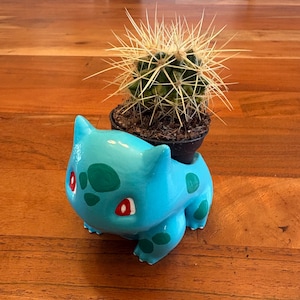 May include: A light blue ceramic planter in the shape of a cartoon character, holding a small potted cactus. The planter has green spots and red accents. The cactus has long, sharp spines.