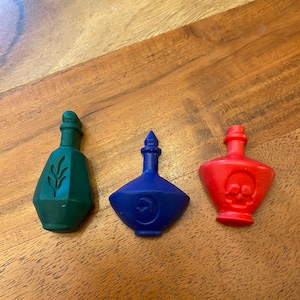 May include: Three small glass bottles with different colored wax. The bottles are shaped like traditional potion bottles. The green bottle has a leaf design, the blue bottle has a crescent moon design, and the red bottle has a skull and crossbones design.