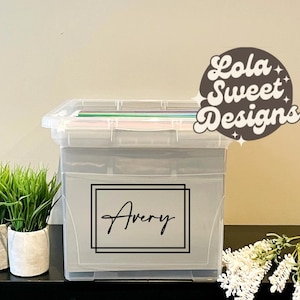 Peut inclure: Une boîte de rangement en plastique transparent avec une étiquette "Avery" dans un cadre noir. La boîte contient des papiers et se trouve à côté de deux petites plantes en pot et d'une branche de fleurs blanches. Le logo Lola Sweet Designs est en haut à droite.