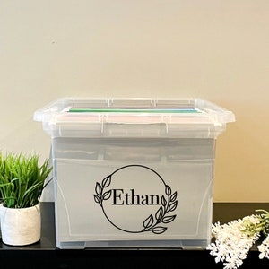 Peut inclure: Une boîte de rangement en plastique transparent avec un autocollant noir et blanc qui dit "Ethan" dans un cercle avec des feuilles autour. La boîte est remplie de dossiers colorés et repose sur une surface noire avec deux plantes en pot.