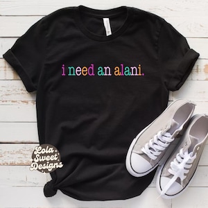 Peut inclure: T-shirt noir avec l'inscription "i need an alani" en couleurs arc-en-ciel. Le t-shirt est associé à des baskets en toile grises avec des lacets blancs. Le logo Lola Sweet Designs est visible sur le t-shirt.