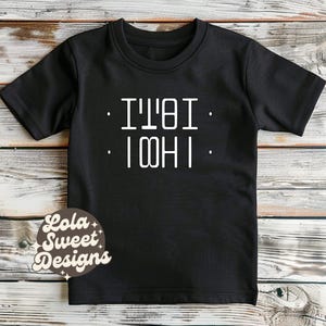 Peut inclure: T-shirt noir à manches courtes avec texte blanc : "I'M. IT'S. I'M HI." Le t-shirt a un col rond et est fait d'une matière douce. Le t-shirt est présenté sur une surface en bois.